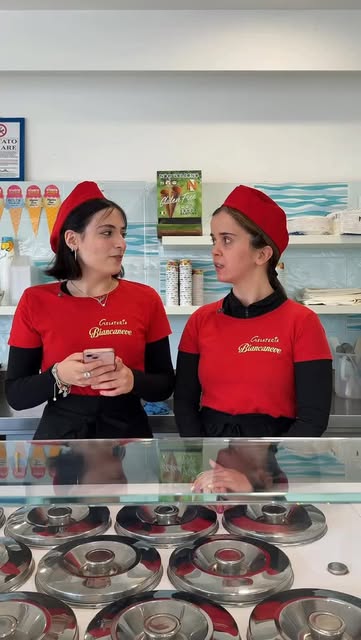 Due gelataie in divisa rossa dietro il banco vetrina della gelateria